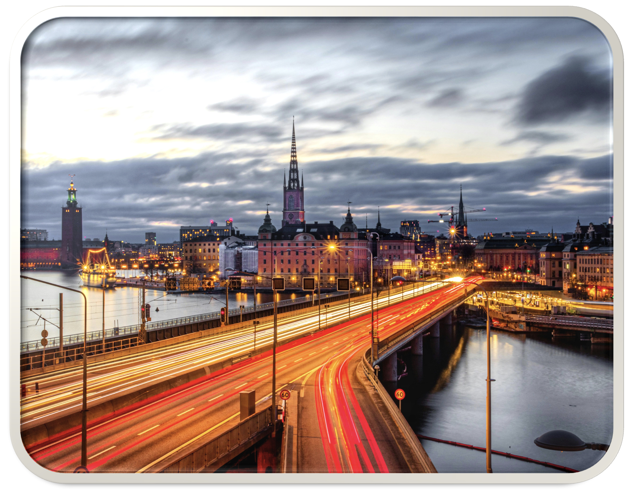 Photographie en pose longue de Stockholm au crépuscule, montrant des traînées lumineuses de la circulation sur le pont Centralbron au premier plan, avec la flèche de l'église de Riddarholmen et l'Hôtel de Ville de Stockholm visibles dans la ligne d'horizon derrière des bâtiments en bord de mer illuminés sous un ciel nuageux dramatique.